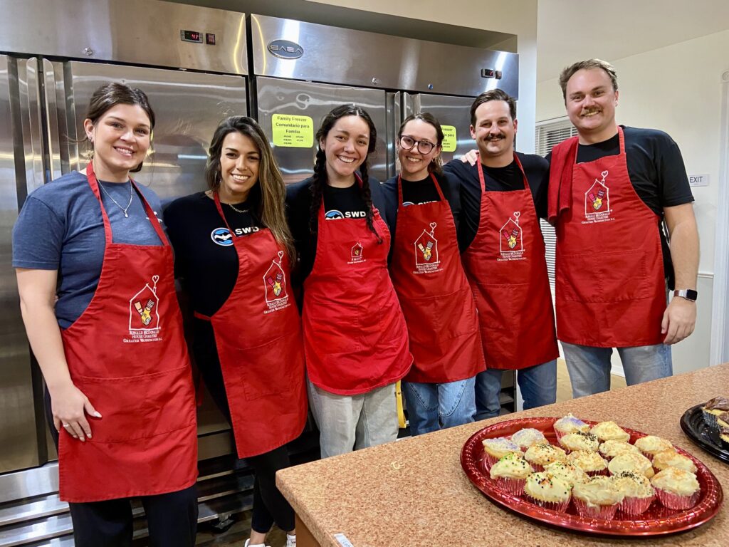 A group of volunteers from Strategic Wealth Designers smile in red Ronald McDonald House aprons beside a tray of muffins in the kitchen as they participated in our Feed the House program