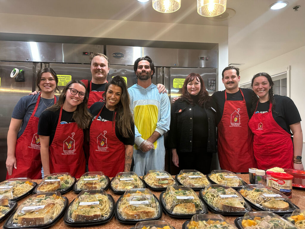 A group of volunteers from Strategic Wealth Designers smile in red Ronald McDonald House aprons along with the Washington Commanders Sam Hartman dressed in costume for Halloween week — while they participated in our Feed the House program