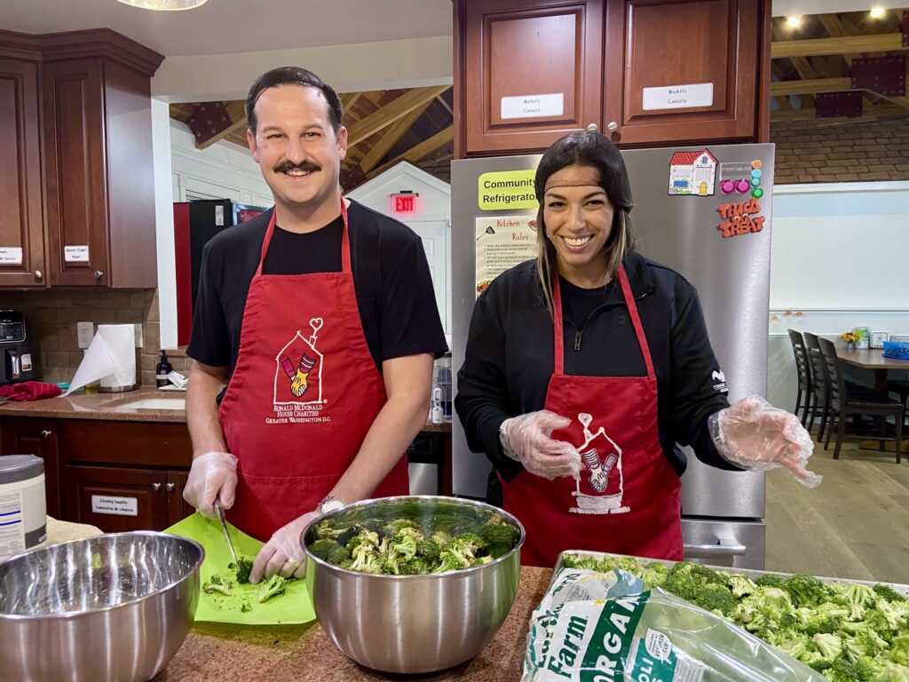 Two volunteers from Strategic Wealth Designers smile in red Ronald McDonald House aprons as they prepare broccoli as part of dinner in the kitchen as they participated in our Feed the House program